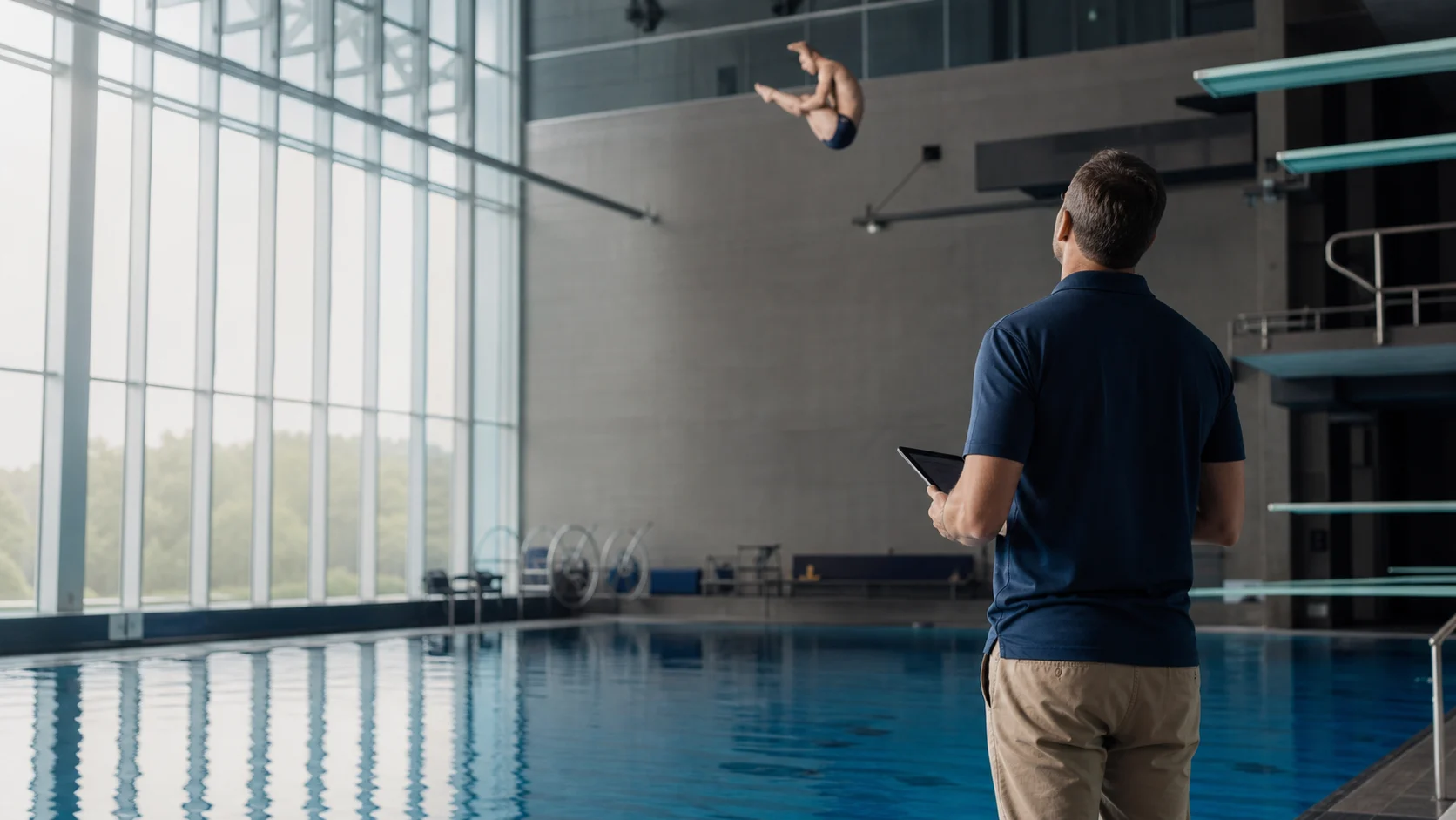 A college diving coach watching a diver practice from the pool deck
